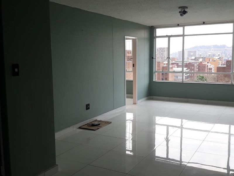 Spacious, empty living area featuring polished white floor tiles, large windows with city views, and neutral green wall paint.