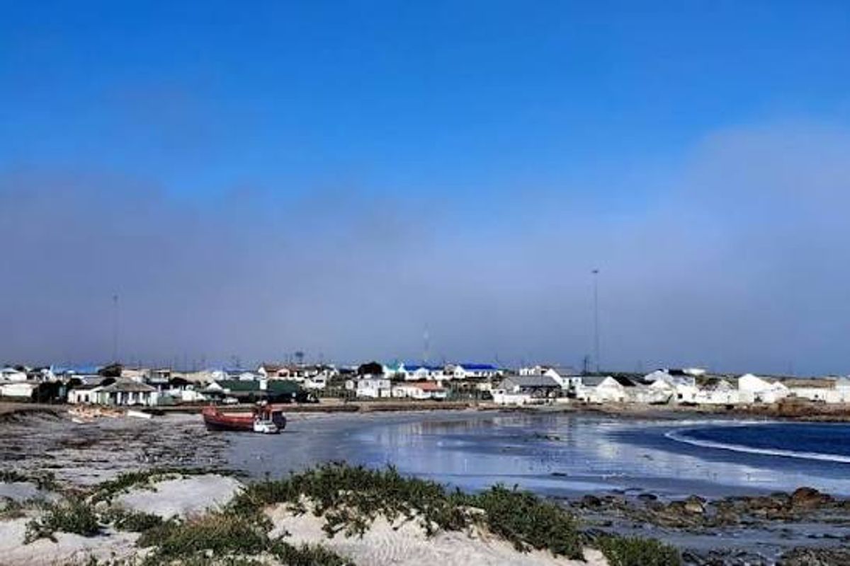 A scenic view of the coastal landscape featuring a sandy beach, rocky shoreline, and distant residential buildings under a clear blue sky.