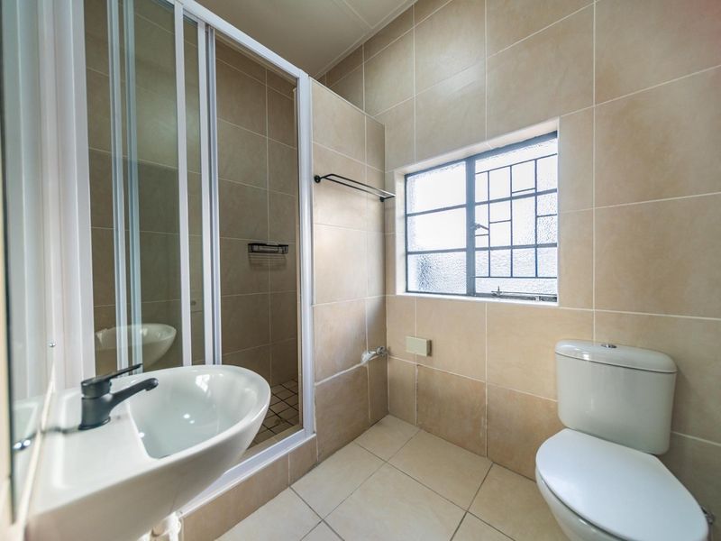 A clean, functional bathroom featuring a shower enclosure, pedestal basin, toilet, and neutral floor-to-ceiling ceramic tiling with a window for natural light.