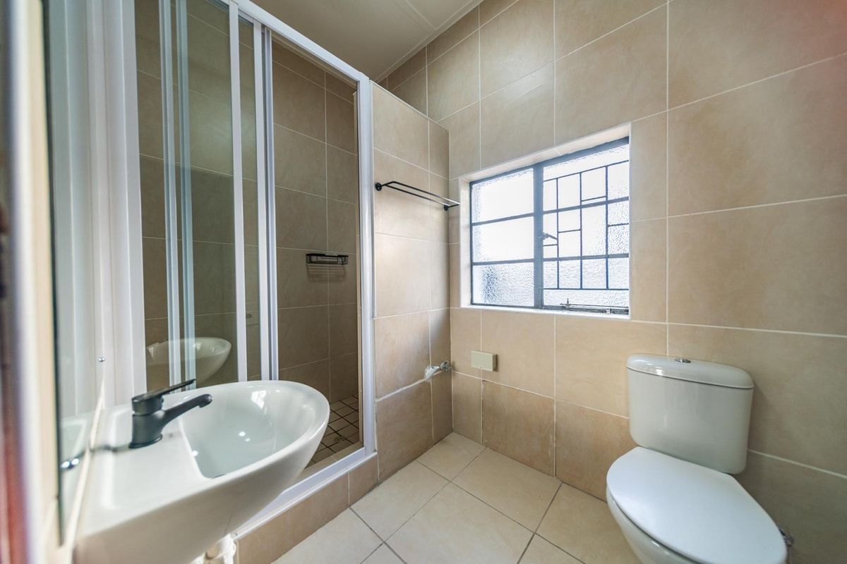A clean, functional bathroom featuring a shower enclosure, pedestal basin, toilet, and neutral floor-to-ceiling ceramic tiling with a window for natural light.