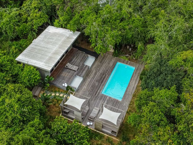 Aerial view of a secluded property featuring a swimming pool, extensive timber decking, and modern outdoor shade structures nestled within a lush forest setting.