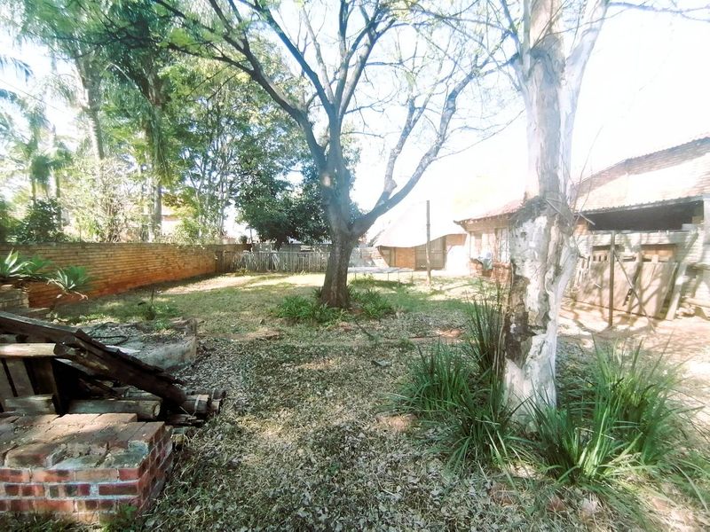 A spacious rear garden featuring mature trees, a brick boundary wall, and a built-in brick fire pit area requiring landscaping and maintenance.