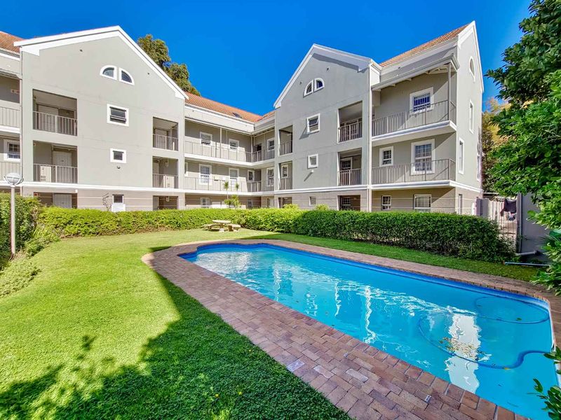 A well-maintained communal swimming pool area featuring a brick-edged pool, manicured lawn, and hedging, set against the backdrop of a multi-story apartment building.