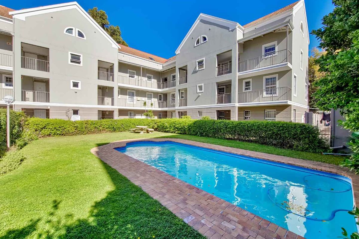 A well-maintained communal swimming pool area featuring a brick-edged pool, manicured lawn, and hedging, set against the backdrop of a multi-story apartment building.