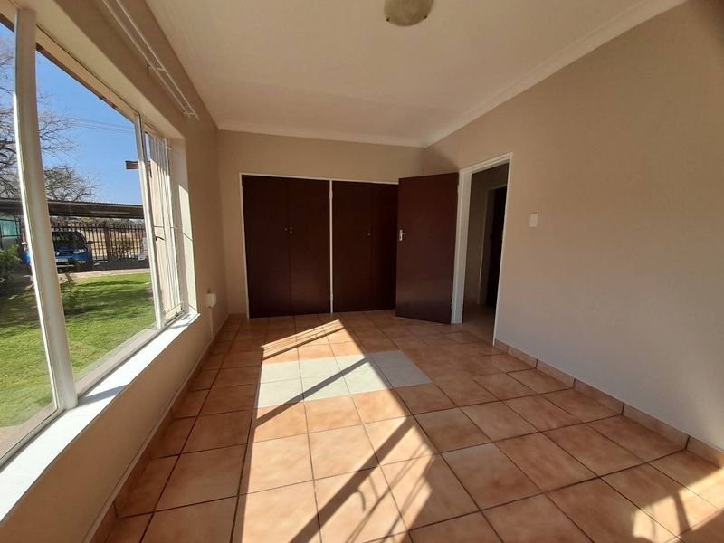 A bright, empty bedroom featuring tiled flooring, built-in cupboards, and large windows providing natural light and garden views.