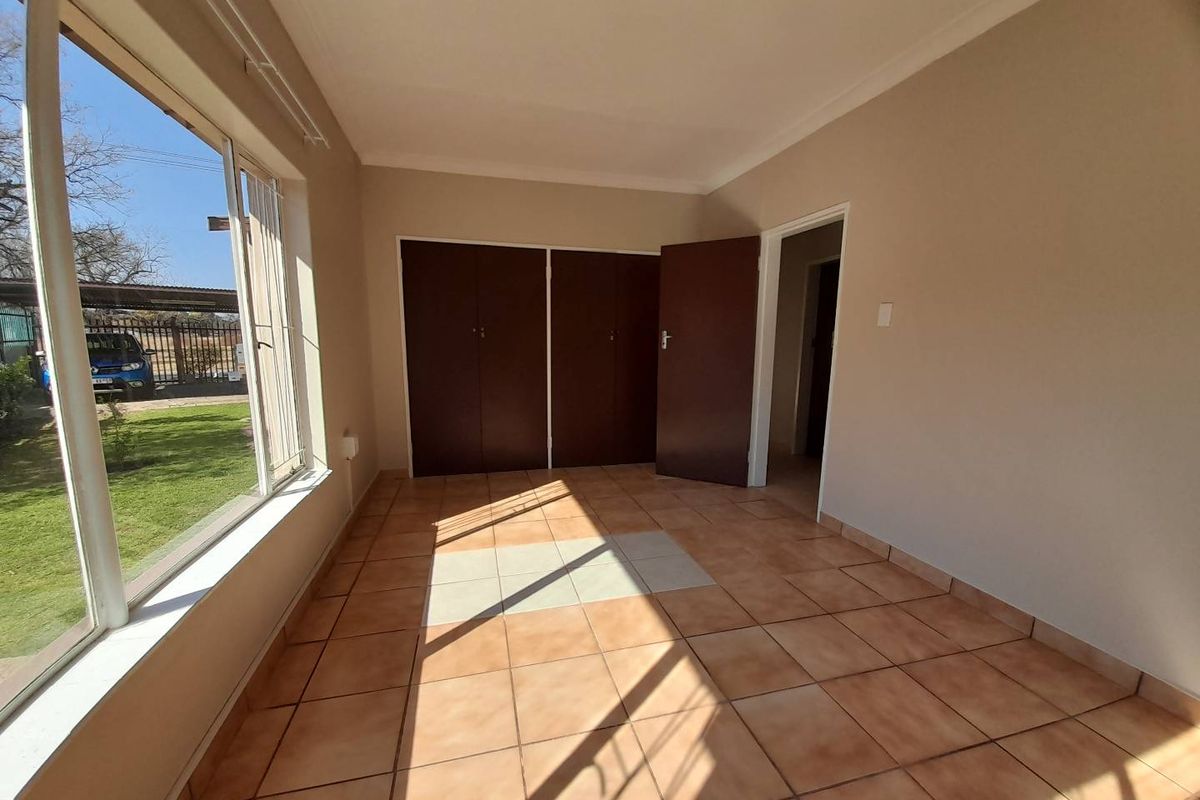 A bright, empty bedroom featuring tiled flooring, built-in cupboards, and large windows providing natural light and garden views.