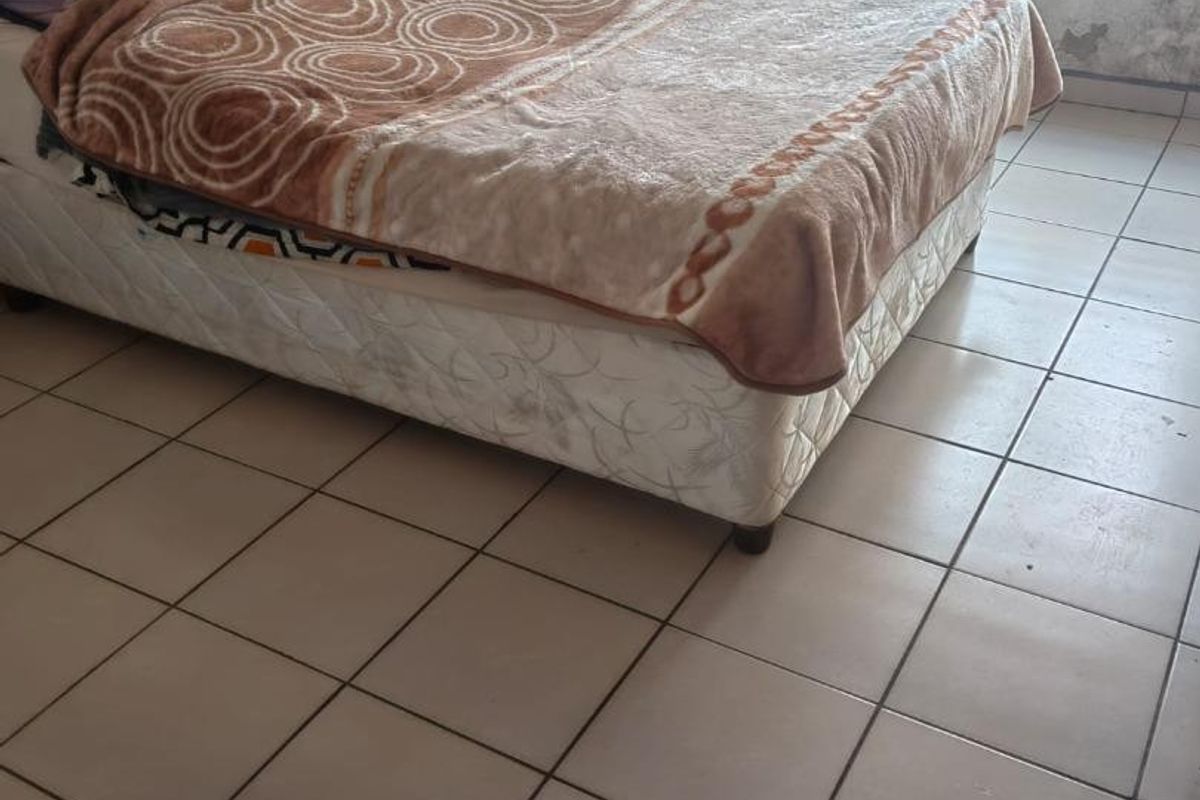 A bedroom featuring tiled flooring and a window with security bars, currently showing significant wall surface deterioration and peeling paint.