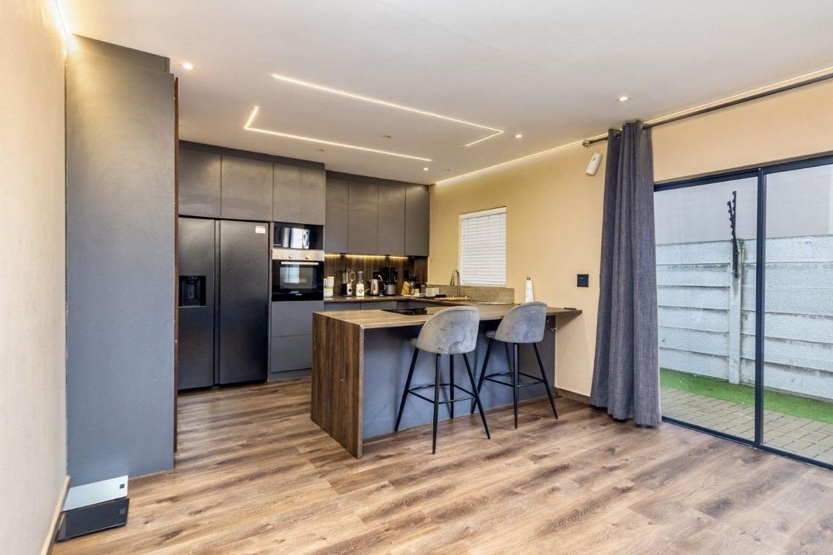 Modern kitchen featuring sleek dark cabinetry, a functional island with breakfast bar seating, integrated appliances, and recessed lighting, opening onto a private outdoor area.