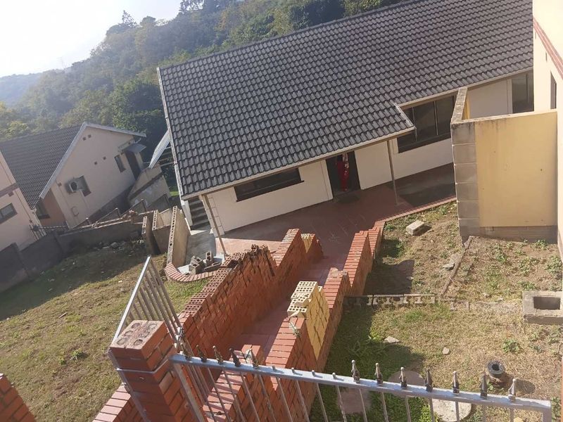 Rear exterior view of the property featuring a tiled roof, paved walkway, and unfinished brick retaining walls on a sloped garden plot.