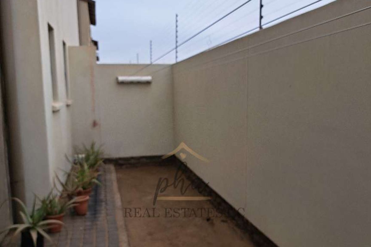 A side exterior area of the townhouse featuring paved walkway, boundary wall with electric fencing, and an unpaved garden section.