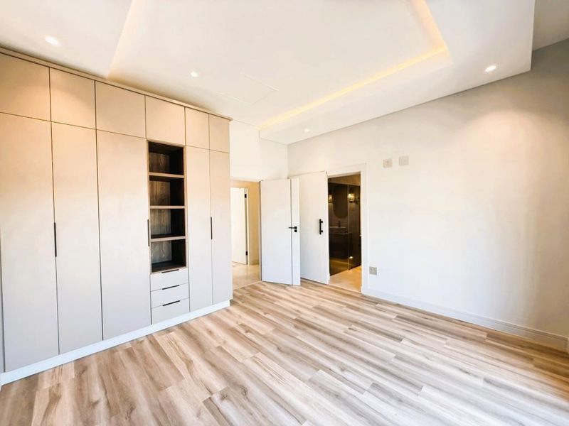 A bright, modern bedroom featuring extensive built-in cabinetry, recessed ceiling lighting, and light wood-look laminate flooring, leading to an en-suite bathroom.