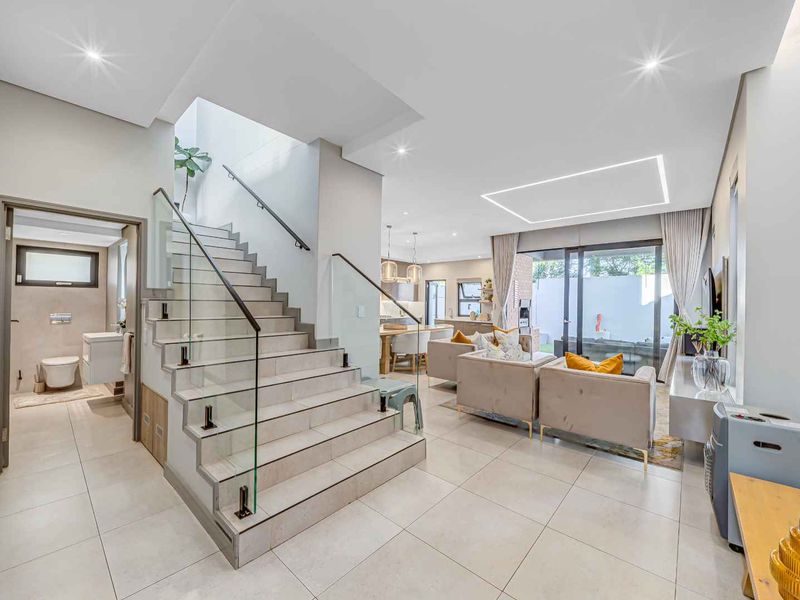 Modern open-plan living area featuring a sleek staircase with glass balustrades, tiled flooring, recessed lighting, and seamless flow to the outdoor patio.