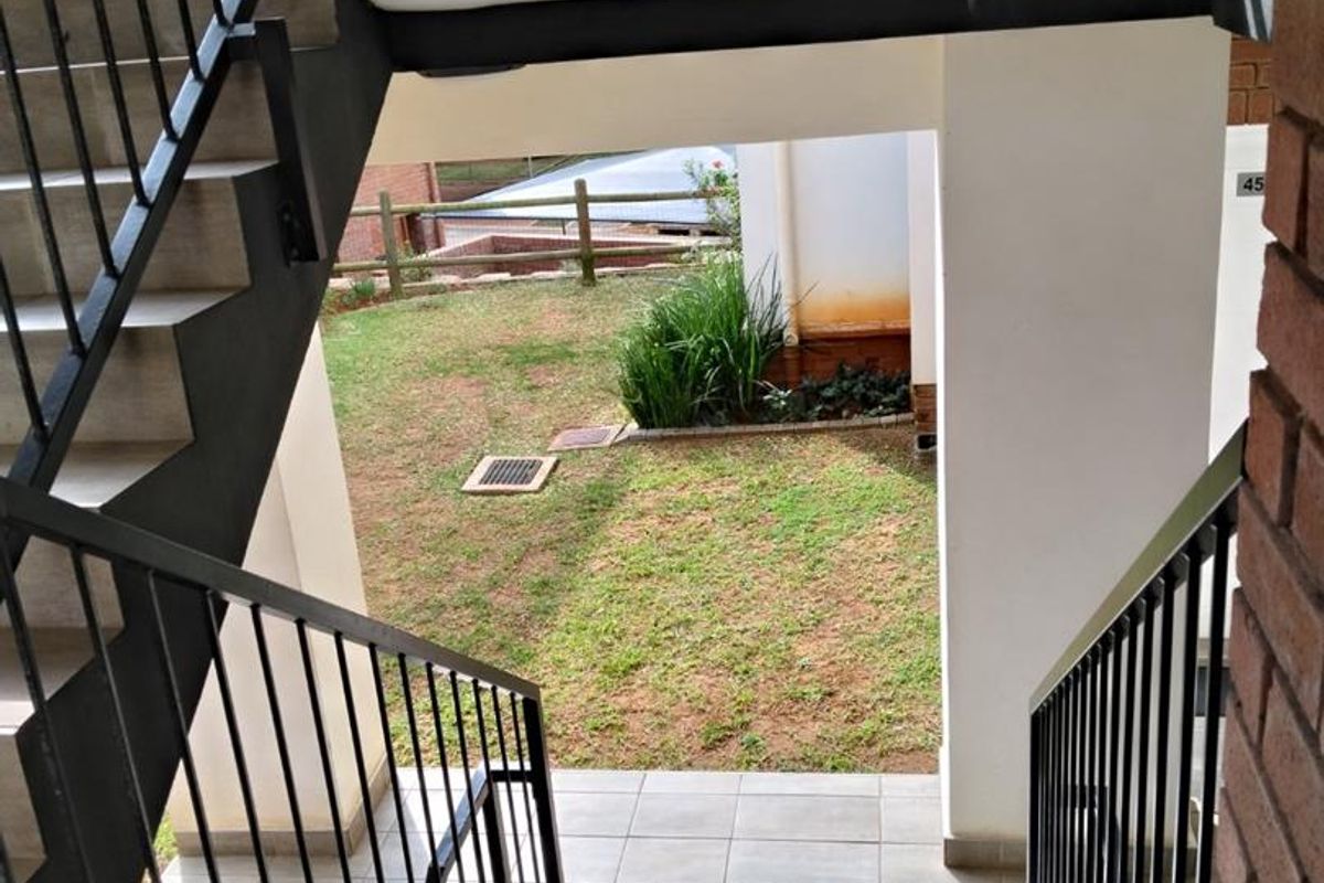 Exterior view looking down a metal staircase towards a communal garden area with tiled landing and brick accents.