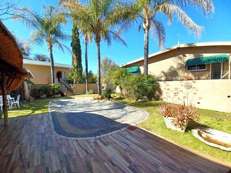 Outdoor pool area featuring a tiled patio, thatched lapa, and landscaped garden with mature palm trees and boundary walling.