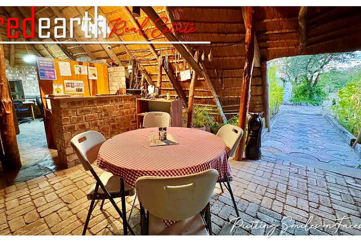 Rustic lodge interior featuring a traditional thatched roof, exposed timber support beams, and a brick-built service counter, opening onto a paved outdoor area.