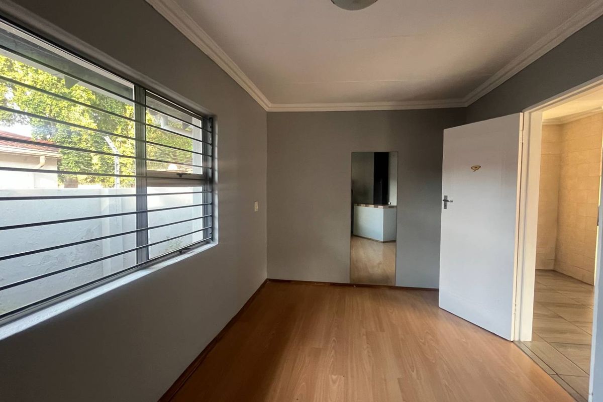 A clean, empty bedroom featuring laminate flooring, security bars on the window, and a built-in wall mirror, leading into an adjacent tiled space.