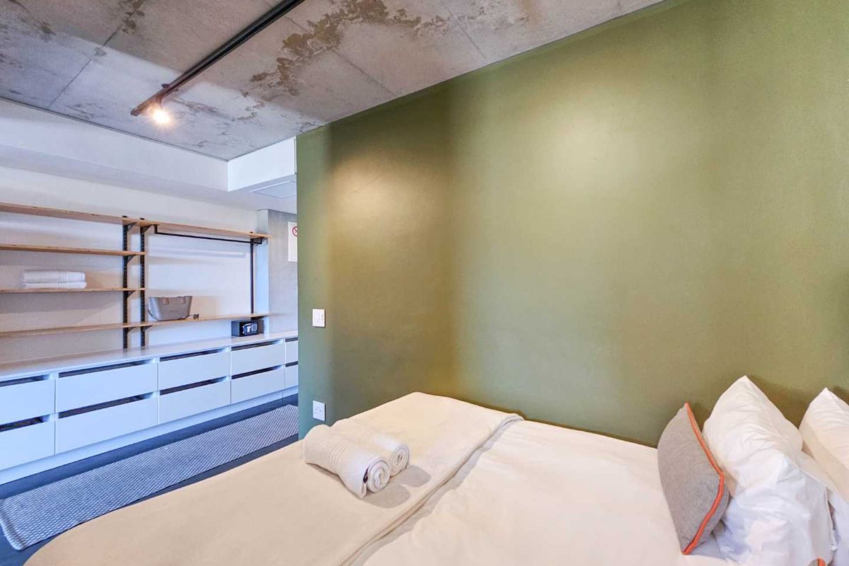 A modern bedroom featuring an industrial-style exposed concrete ceiling, olive green accent wall, and built-in white cabinetry with open shelving.