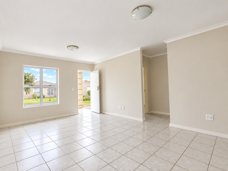 A bright, empty living area featuring neutral tiled flooring, coved ceilings, and direct access to the exterior through a glass-paneled door.