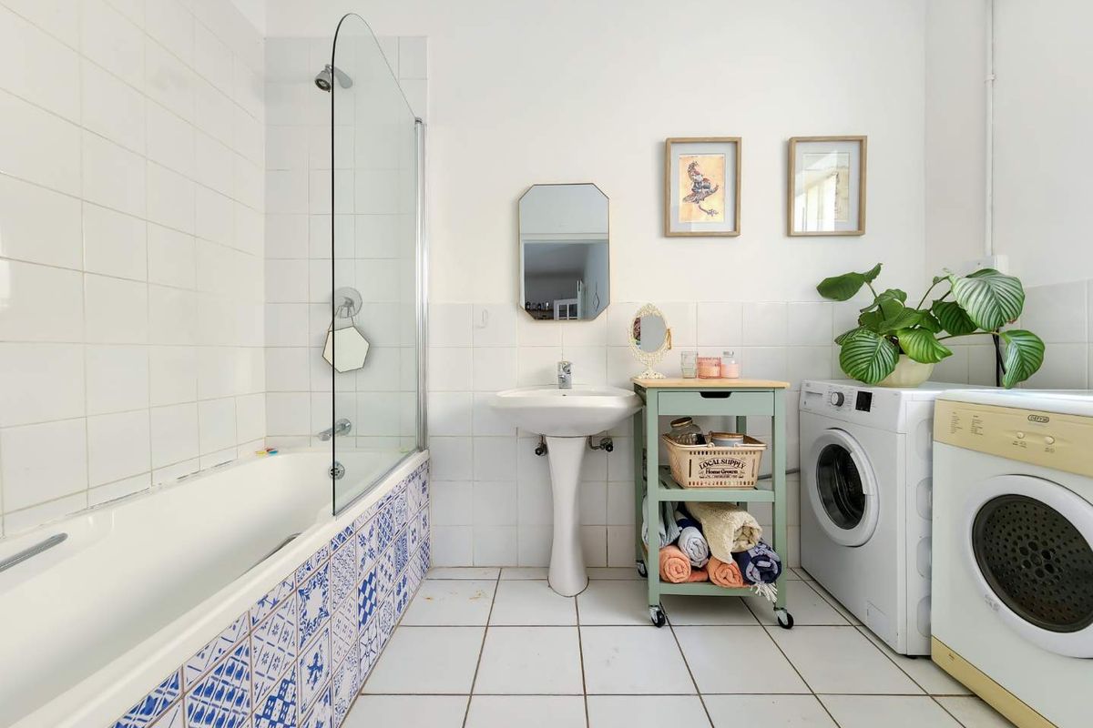 A functional bathroom featuring a pedestal basin, bathtub with glass screen, and tiled finishes, with integrated space for laundry appliances.