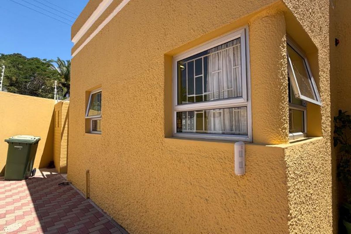 A side view of the property featuring textured yellow exterior walls, aluminium window frames with security bars, and a paved walkway.