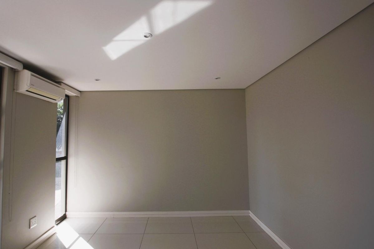 A bright, empty bedroom featuring neutral wall tones, tiled flooring, recessed ceiling lighting, and a wall-mounted air conditioning unit.