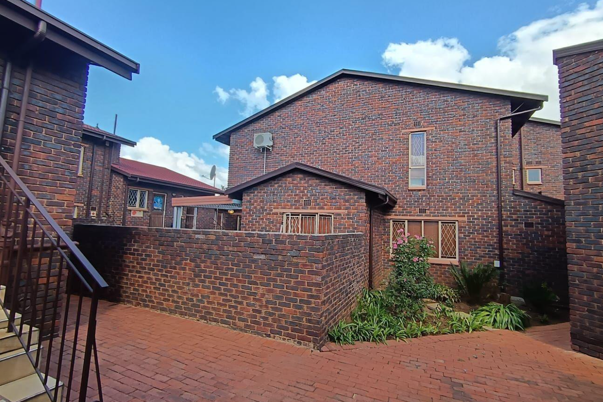 A rear view of a face brick cluster home featuring a paved courtyard, security-fitted windows, and an external air conditioning unit.