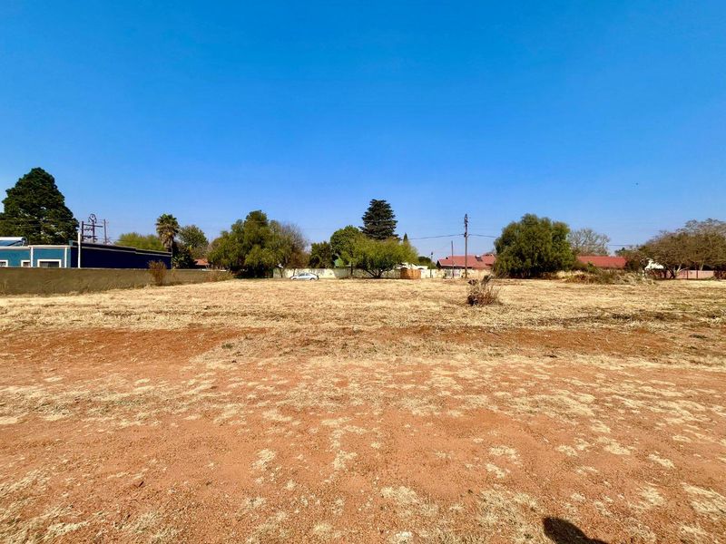 A vacant residential plot featuring dry grass, mature trees, and a perimeter wall, offering a clear space for future development.