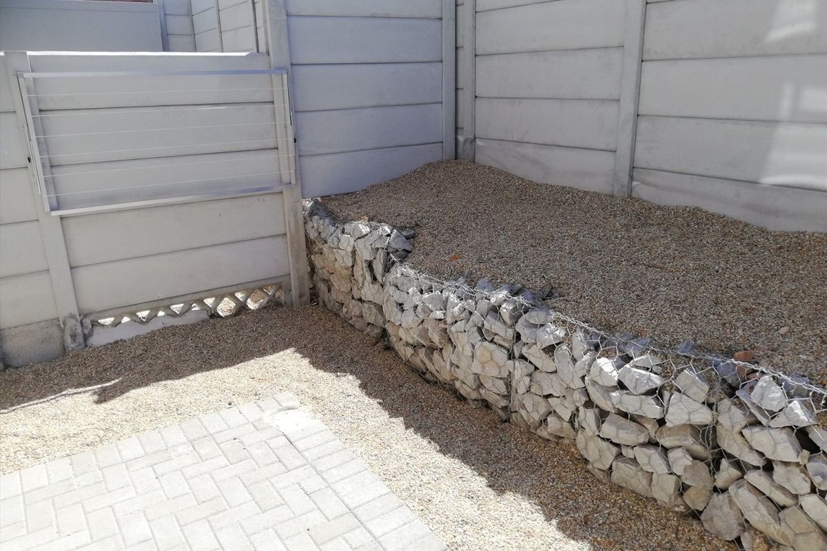 A rear courtyard area featuring a gabion retaining wall, gravel landscaping, paved sections, and a precast concrete boundary wall with a mounted drying rack.