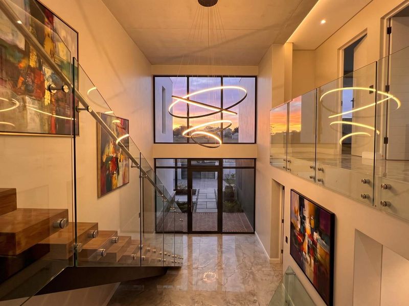 Grand double-volume entrance hall featuring a striking timber staircase, glass balustrades, marble flooring, and a contemporary chandelier with expansive window views.
