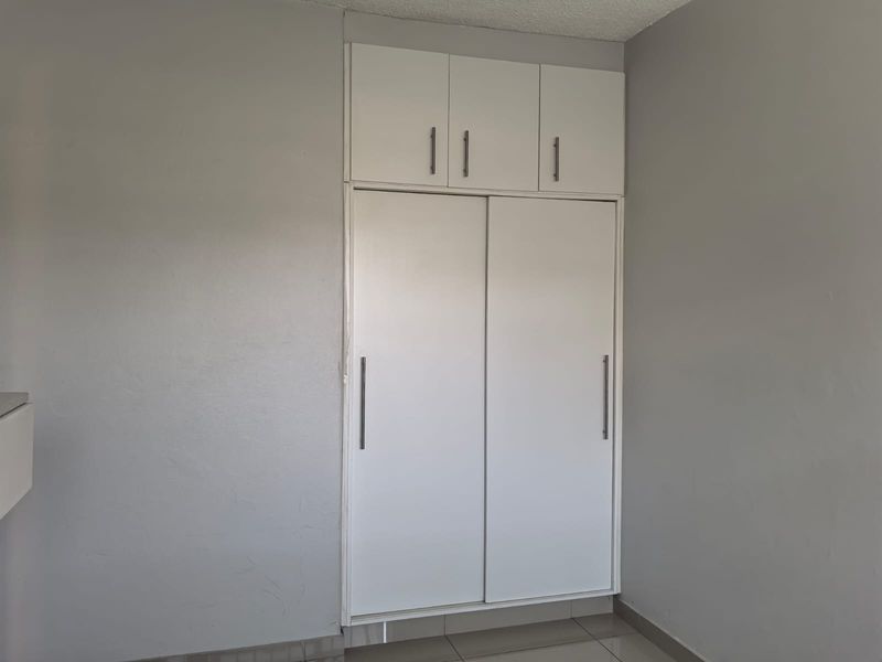 A clean bedroom interior featuring a white built-in wardrobe with overhead storage cupboards and neutral tiled flooring.