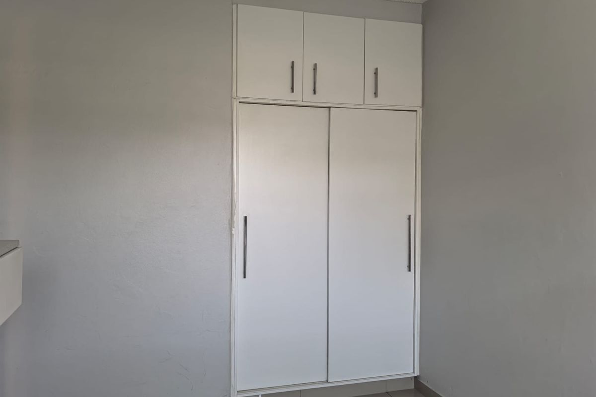 A clean bedroom interior featuring a white built-in wardrobe with overhead storage cupboards and neutral tiled flooring.
