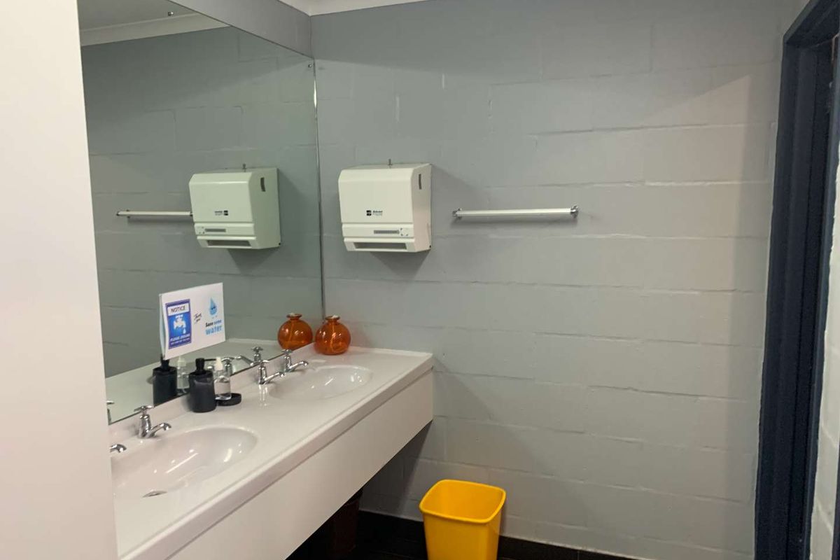 A functional office restroom featuring a dual-basin vanity, large wall-mounted mirror, and paper towel dispenser against painted blockwork walls.