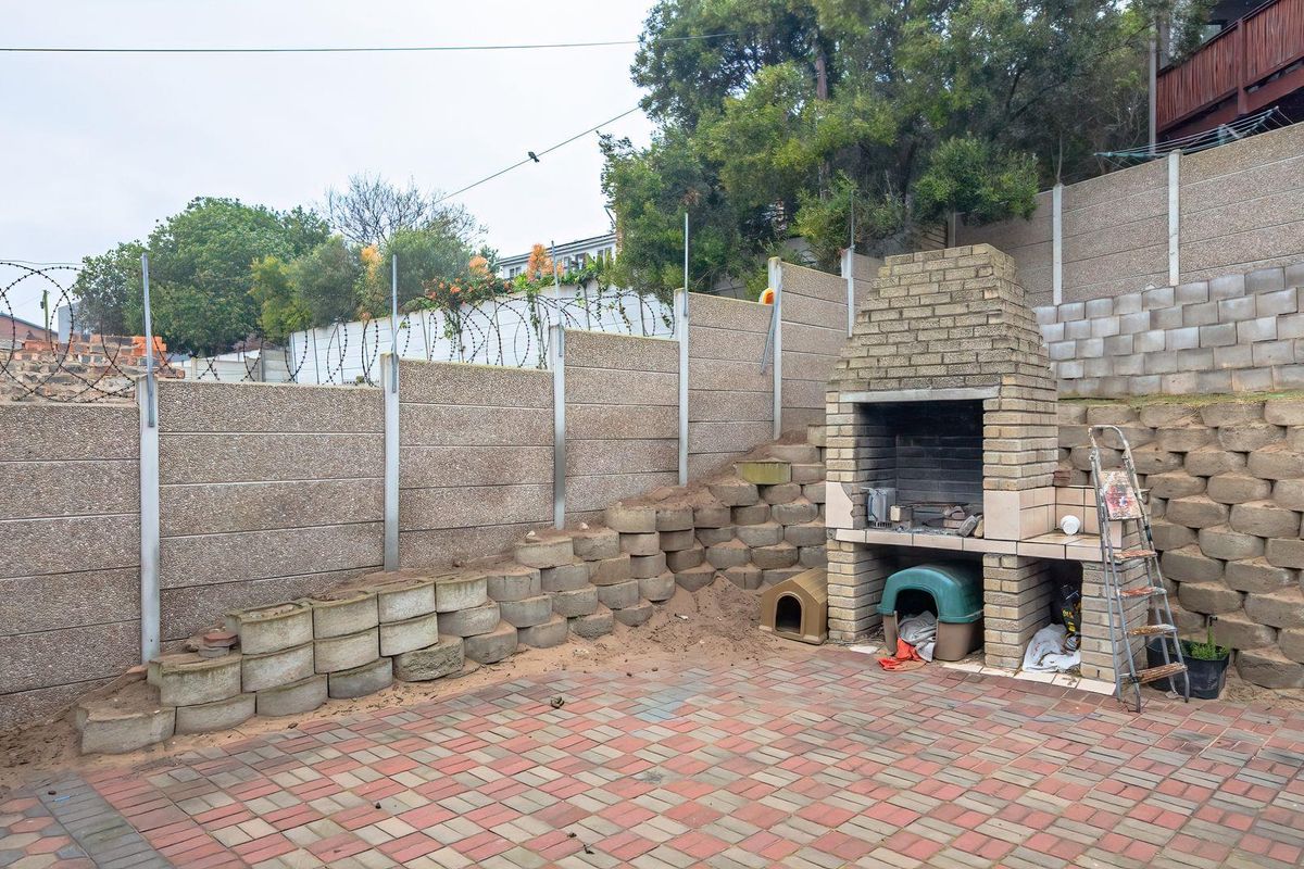 Rear outdoor area featuring a built-in brick braai, paved patio, and tiered retaining walls with perimeter security fencing.