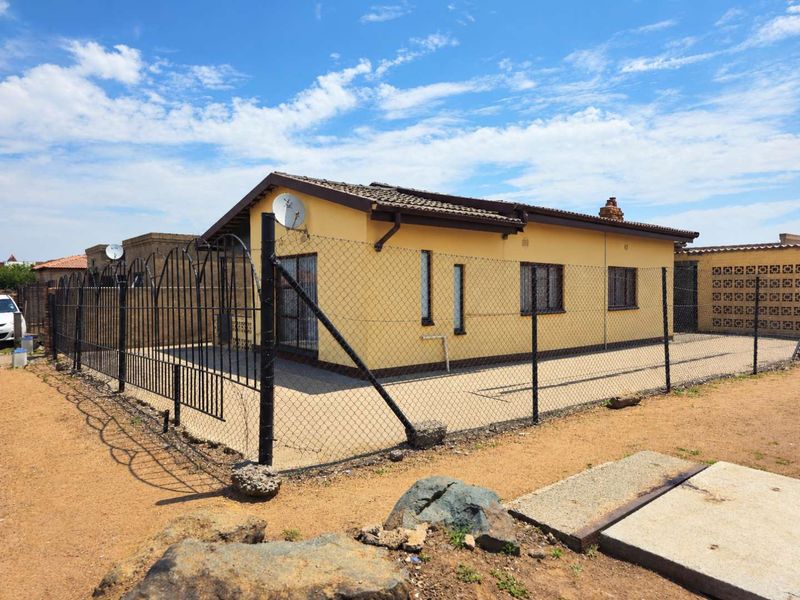 A detached single-story house featuring a tiled roof, yellow exterior walls, and a secure perimeter fence with a gated entrance.