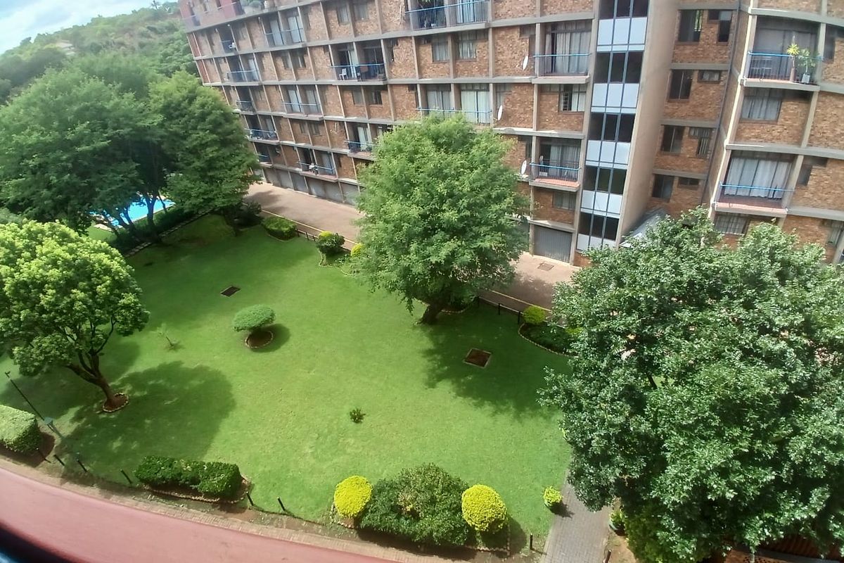 Elevated view overlooking the apartment complex grounds, featuring a well-maintained communal garden, mature trees, paved pathways, and a swimming pool area.