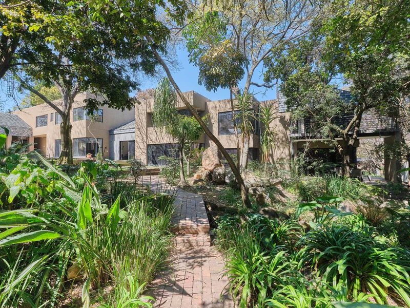 A view of the rear building facade featuring a brick pathway leading through a lush, mature garden with established trees and a balcony.