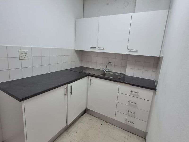 A compact office kitchenette featuring white cabinetry, dark laminate countertops, a stainless steel sink, and tiled backsplash in a functional, utilitarian space.