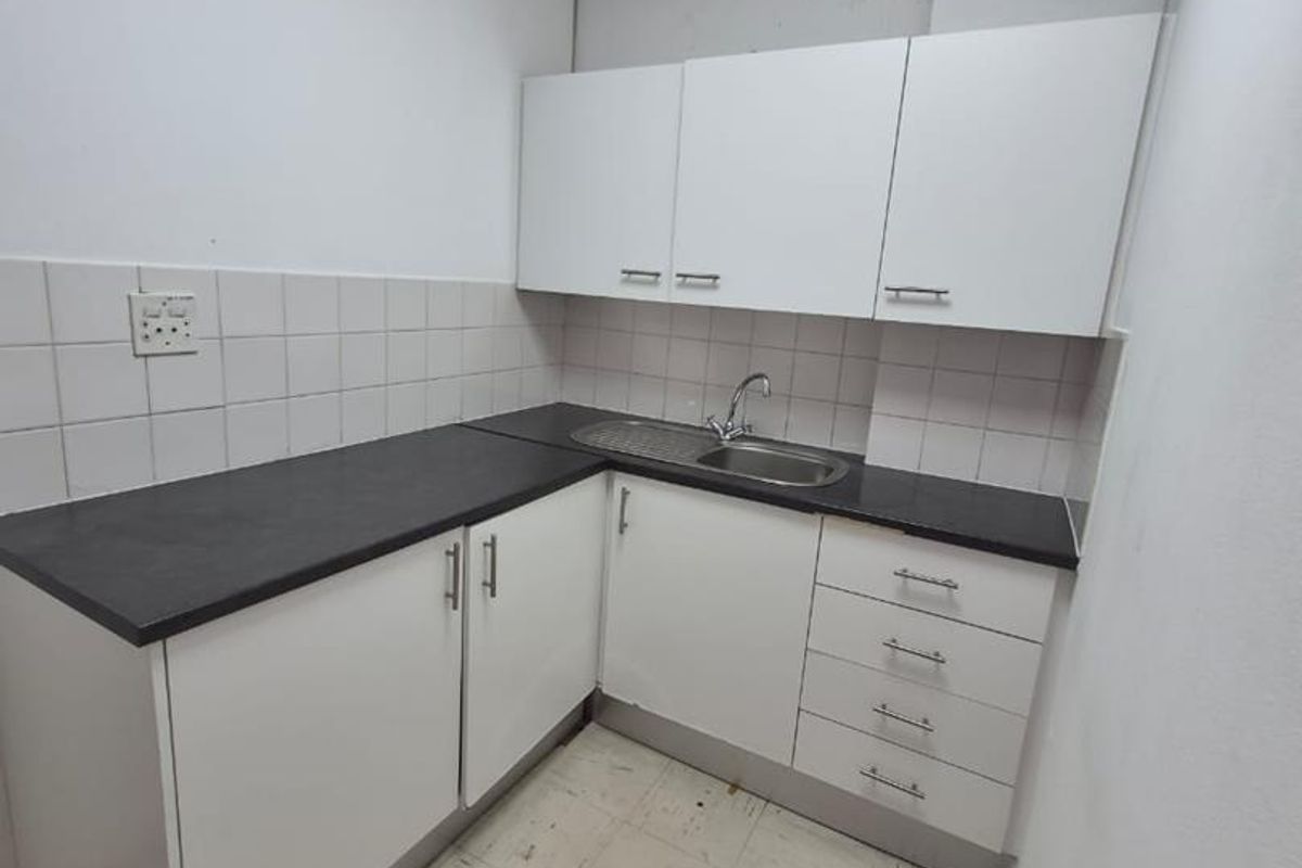A compact office kitchenette featuring white cabinetry, dark laminate countertops, a stainless steel sink, and tiled backsplash in a functional, utilitarian space.