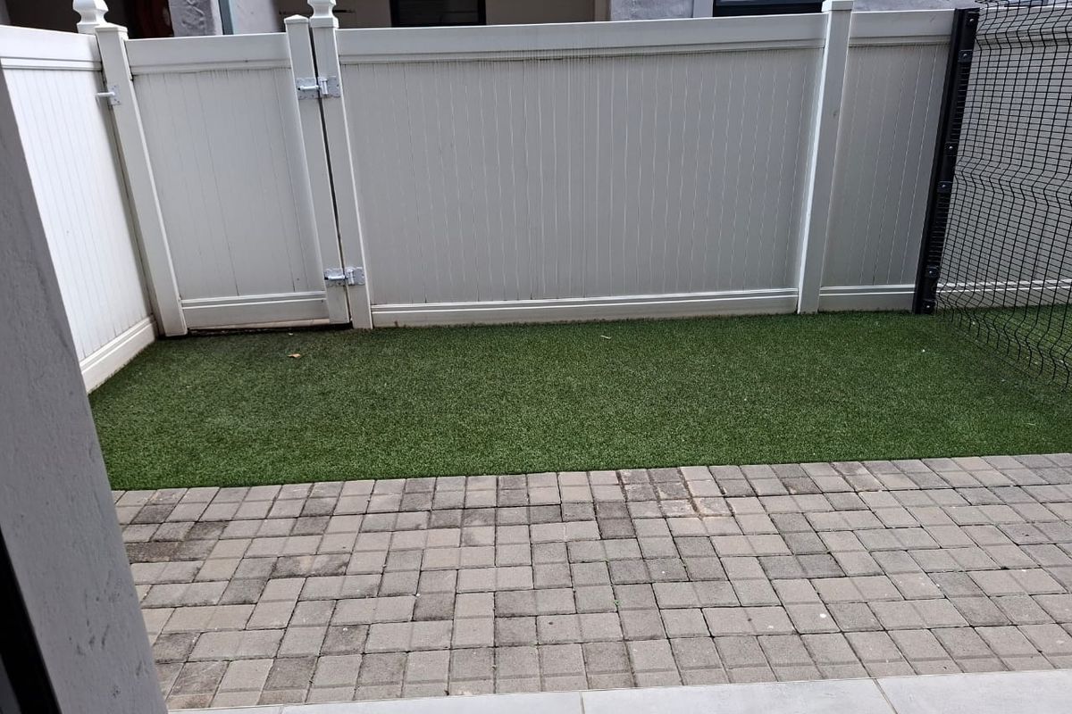 A private rear courtyard featuring a paved patio area, artificial turf lawn, and a white vinyl privacy fence with a gate.