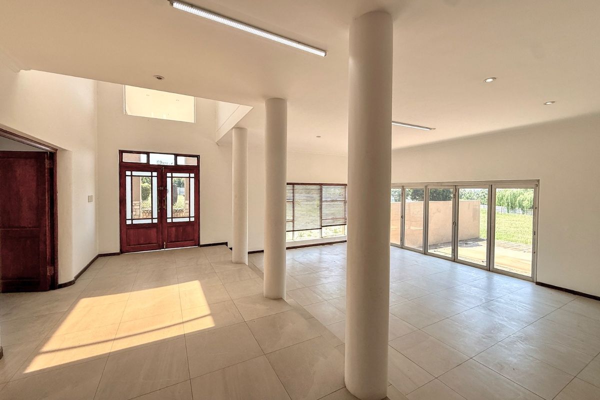 Spacious, open-plan living area featuring large structural columns, neutral tiled flooring, and abundant natural light through double entrance doors and sliding glass patio access.