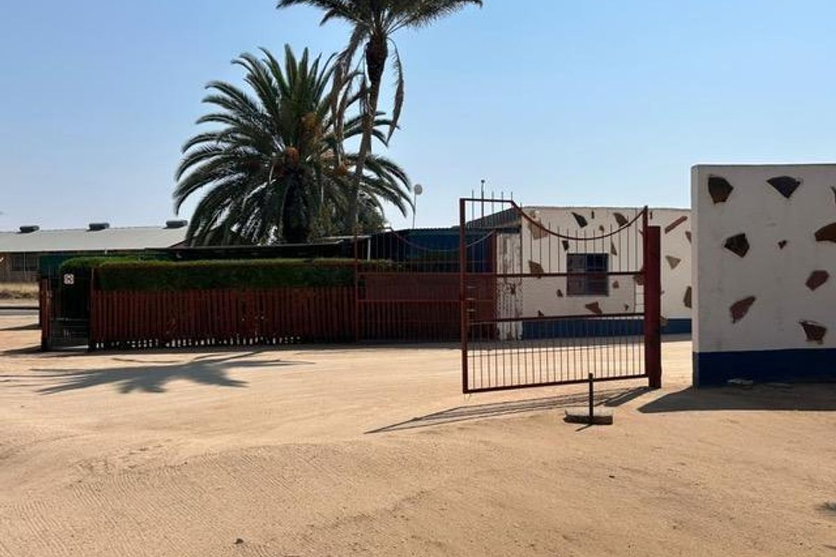 Front exterior view of the industrial property featuring a metal security gate, perimeter fencing, and a large unpaved driveway area.