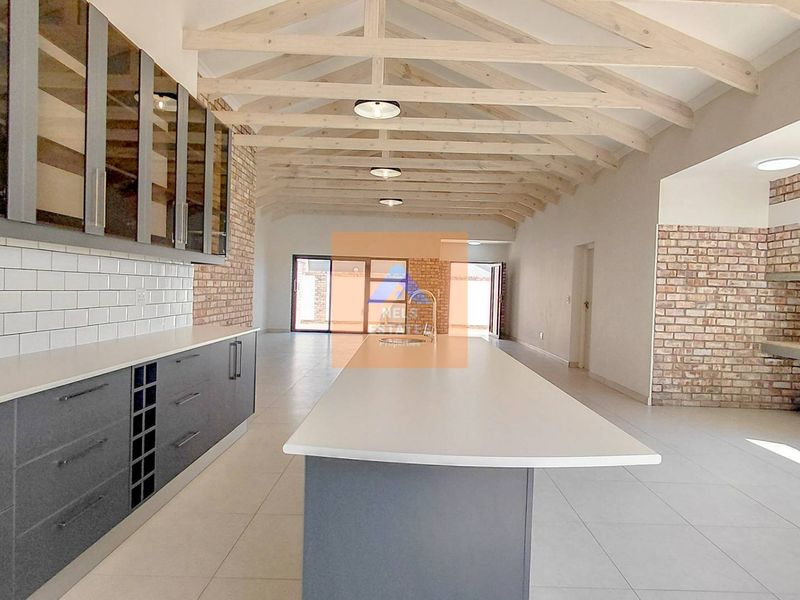 Spacious open-plan kitchen and living area featuring exposed timber trusses, a large central island, modern cabinetry, and a rustic brick fireplace.