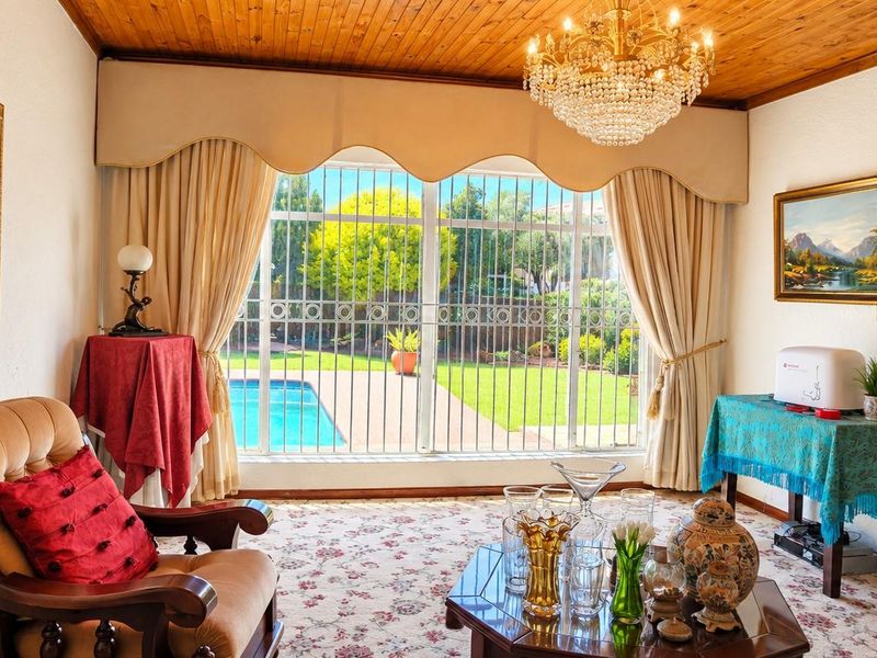 A living room featuring a timber ceiling, crystal chandelier, and a brick archway, with large windows overlooking the garden and swimming pool.