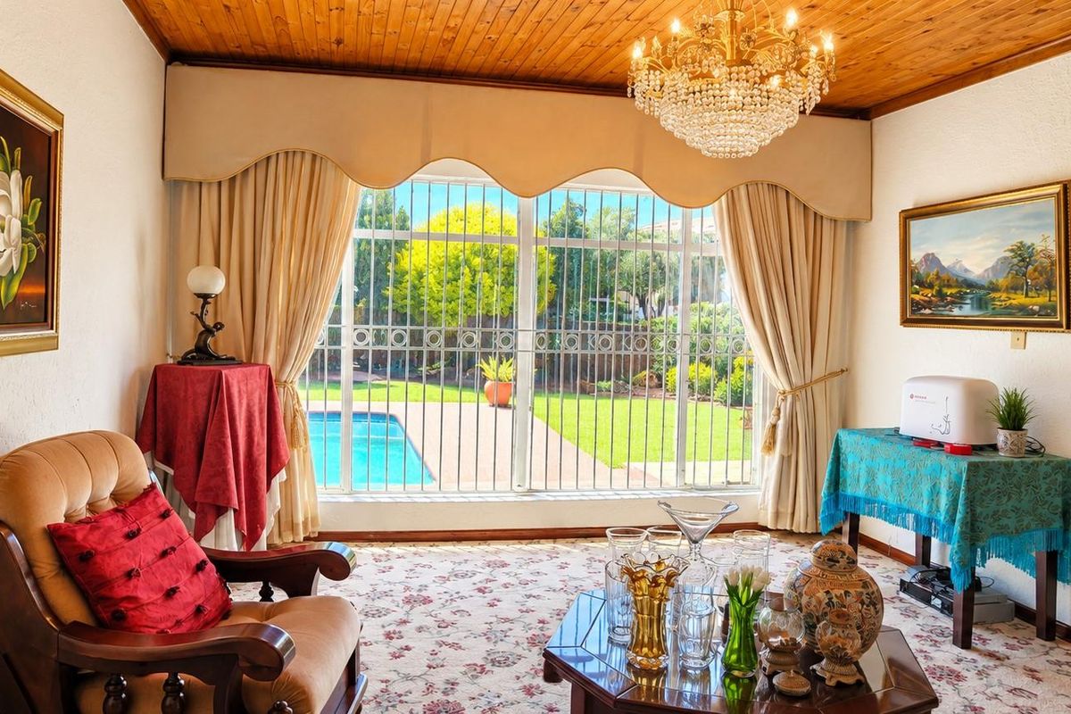 A living room featuring a timber ceiling, crystal chandelier, and a brick archway, with large windows overlooking the garden and swimming pool.