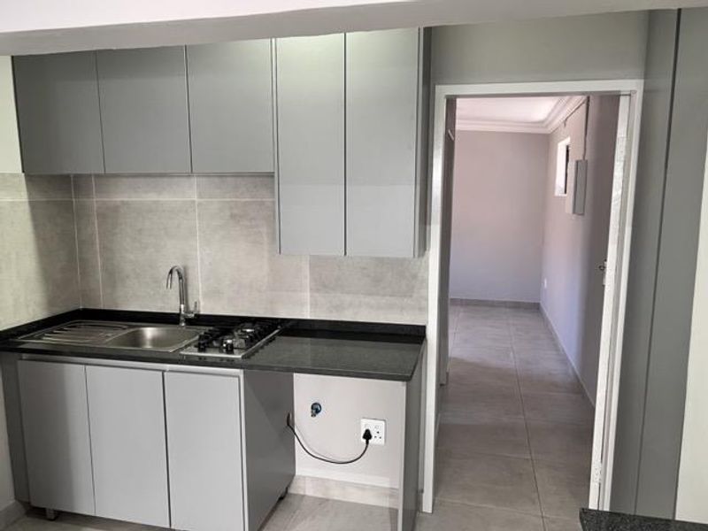 A modern, compact kitchen featuring grey cabinetry, stone countertops, a gas hob, and a stainless steel sink, with neutral tiled flooring throughout.
