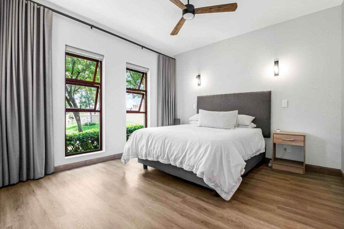 A bright and modern bedroom featuring wood-look flooring, large windows with curtains, a ceiling fan, and wall-mounted lighting fixtures.