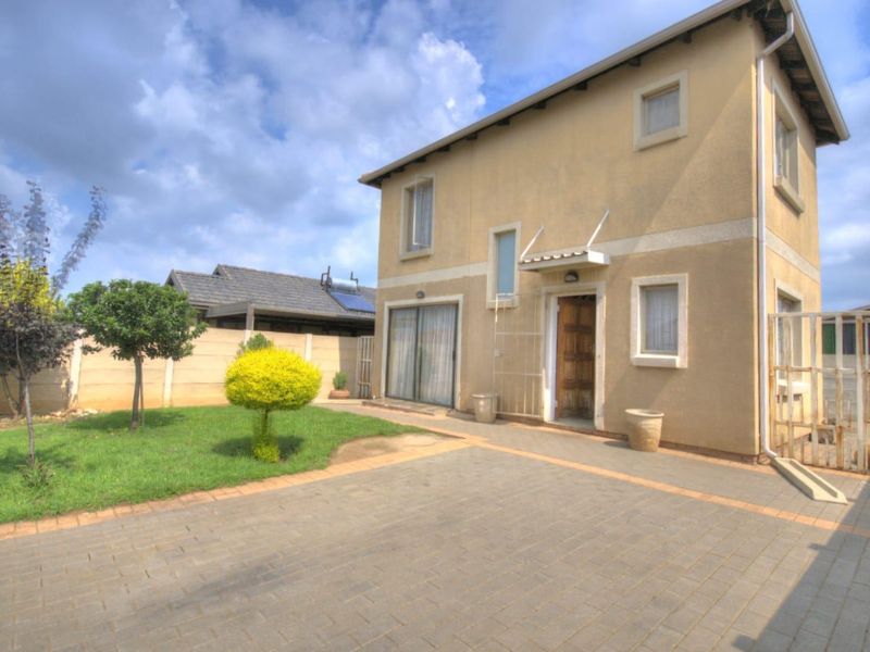 A rear exterior view of a two-story house featuring a paved driveway, a small lawn, and secure boundary walls.