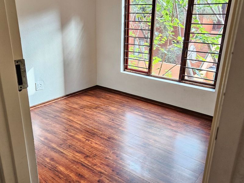 A bright, empty bedroom featuring warm-toned laminate flooring and a large window fitted with security burglar bars.