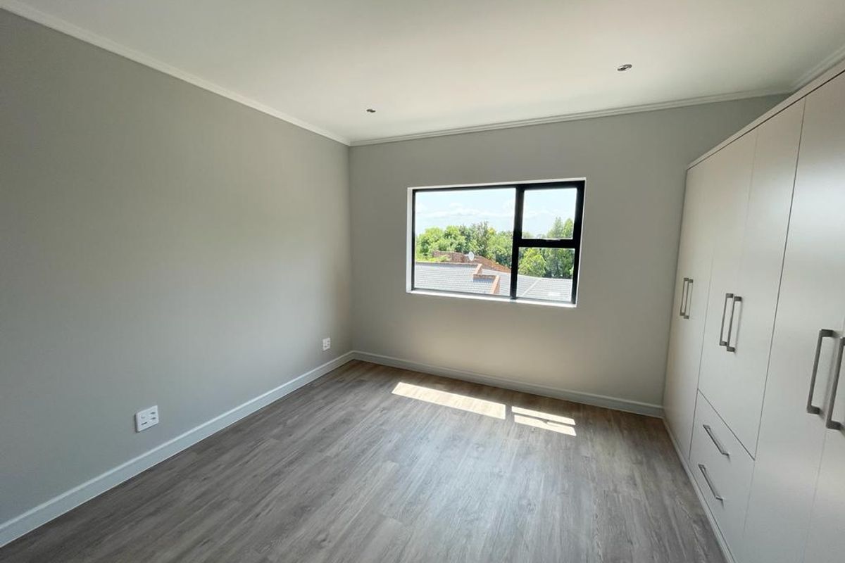 A bright, empty bedroom featuring modern laminate flooring, neutral grey walls, and ample built-in cupboard storage with a view through the window.