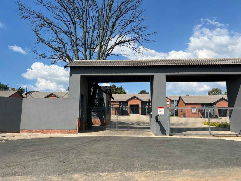 Front entrance view of the apartment complex featuring a secure gated access point, paved driveway, and perimeter walling with electric fencing.